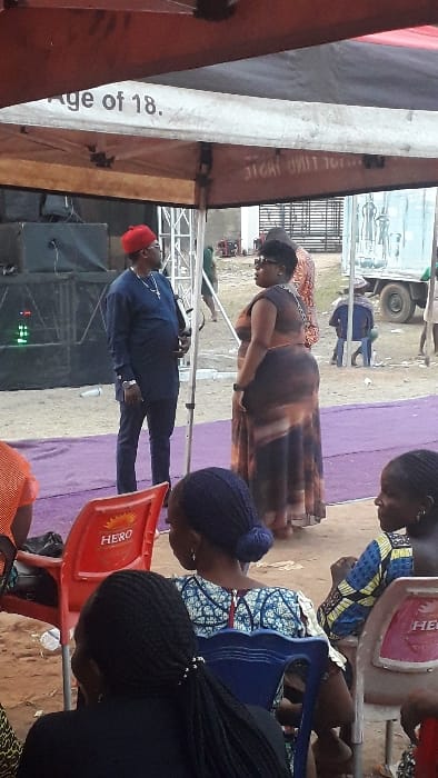 The scene depicts an outdoor gathering or event in Agukwu Nri, Nigeria. A white canopy with a red trim partially covers the top of the frame, displaying text "Age of 18." and inverted text "JI GAI TINI TILL..." on its underside. The ground is an uneven reddish-brown dirt, with a purple mat laid across portions of it, including under the main subjects.
In the midground, a man and a woman stand facing each other, engaged in conversation. The man on the left wears a navy blue long-sleeved tunic and trousers, a red cap, dark sunglasses, and a silver chain with a cross pendant. He has his hands clasped in front of him. The woman on the right wears a sleeveless, form-fitting long dress with a brown and orange/tan tie-dye pattern. She has dark, styled hair, dark sunglasses, and a white strap visible over her right shoulder.
Behind them, on the left, is a dark-colored stage structure with large speakers or equipment and metal trussing. Green lights are visible on one piece of equipment on the stage.
In the foreground, partially obscured, are several individuals seated with their backs to the camera. One woman on the left wears orange and is seated in a red plastic chair branded with "HERO". Another woman with dark braided hair in a bun wears a blue and white patterned top and sits in a blue plastic chair. A third woman with dark braided hair in a ponytail wears a black top and sits in a red "HERO" branded plastic chair. A fourth woman, further right, is partially visible wearing a yellow, blue, and white patterned top, also seated in a "HERO" branded chair.
In the background, beyond the standing individuals, a light-colored truck is visible on the far right. A fence or gate and parts of structures are also visible. Other indistinct figures are present further in the background, including one person seated on a low stool near the truck.