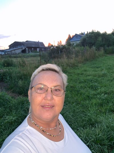 In Суроватиха, Russia, a self-portrait captures a woman with short blonde hair, wearing glasses and a white t-shirt adorned with a beaded necklace. She stands in a grassy field with several rustic houses visible in the background. The sky above is a bright white, suggesting either early morning or late evening light. The overall scene depicts a rural setting with a focus on the woman as the central subject.