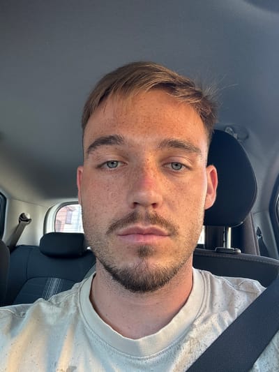 A man is taking a selfie in the driver's seat of a car. The man has fair skin, light brown hair, and blue eyes. He is wearing a light-colored t-shirt and has a beard and mustache. The car's interior is visible behind him, including the headrest and seatbelt. The man appears to be in Gradezhnitsa, Bulgaria, as suggested by the context.