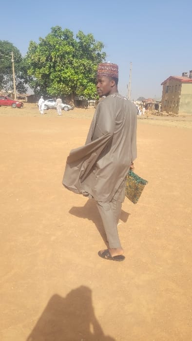 An outdoor scene in the city of Badiko, Nigeria, shows a man in the foreground walking across a barren, reddish-brown dirt ground. The man is observed from behind, turning his head to look over his left shoulder towards the left of the frame. He wears a grey-brown long-sleeved tunic over matching trousers and a patterned, multi-colored cap. Dark sandals are on his feet. In his left hand, he carries a small, textured bag or pouch with green and gold hues. A long shadow of the man and the photographer is cast on the ground in front of him.
In the mid-ground to the left, a large green-leafed tree stands prominently. Behind the tree and slightly to its right, a red vehicle is partially visible. Further back, a group of approximately three individuals, dressed in light-colored attire, are gathered near a silver vehicle. A utility pole is also present in the left background. To the right of the frame, a multi-story building with reddish-brown walls and a darker roof is visible, with other indistinct structures further right. The sky overhead is clear and light blue. The scene is illuminated by bright sunlight.