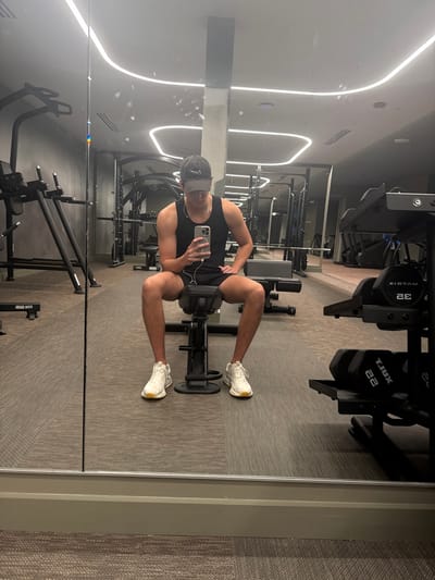 A man in athletic attire sits on a gym weight machine, facing a mirror. He's wearing a tank top, shorts, sneakers, and a baseball cap, holding a phone in his hand. The gym setting features various weight machines and equipment, with neutral-toned walls and flooring. Reflective lights illuminate the ceiling, and the image appears to be a self-portrait taken in the gym.
