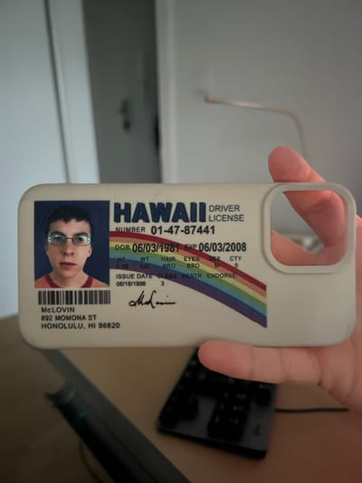 An identity card, designed to resemble a Hawaii driver's license, is held by a person's hand, obscuring a portion of the lower right of the card and the right edge of the card. The identity card features a photograph of a young white male wearing glasses and an orange t-shirt. The text on the card indicates "McLovin" as the name, "892 Momona St, Honolulu, HI 96820" as the address, and includes details such as date of birth, weight, height, eye color, and expiration date. A rainbow stripe is visible on the lower part of the card. The background is out of focus, showing a blurred indoor environment with what appears to be a desk and a keyboard in the foreground. There is no information within the image to suggest a connection to the city of Verden (Aller), Germany.