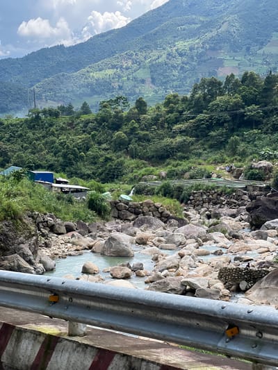 This is a medium shot, eye-level, outdoor photograph taken in Bình Lư, Vietnam. The scene depicts a rocky riverbed with clear, shallow water flowing through it. Large, rounded boulders of varying sizes are scattered throughout the river and along its banks. Lush green vegetation, including trees and grasses, grows abundantly on the surrounding hillsides. In the middle ground, there are several man-made structures, including buildings with blue and green roofs and a series of outdoor fish ponds. A metal guardrail is visible in the foreground, separating the viewer from the river. In the background, majestic, tree-covered mountains rise up, their slopes dotted with patches of cultivation. The sky is partly cloudy, with white clouds against a blue backdrop. The overall impression is one of a natural landscape enhanced by human presence.