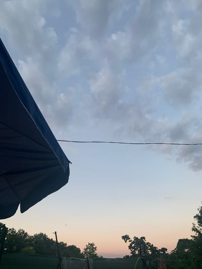The image depicts an outdoor scene at dusk, with a large dark blue object, possibly a tent or canopy, dominating the lower left portion of the frame. This object has a defined edge and a visible seam, suggesting it is a manufactured item. A thin, dark wire stretches diagonally across the sky from the upper left to the mid-right of the image, appearing to be a power line or clothesline.

The sky is a prominent feature, transitioning from a cloudy, greyish-blue on the left to a clear, pale peach hue on the right. The clouds are scattered and textured, indicating a possibly overcast or changing weather condition earlier in the day. The warm tones of the sky on the right suggest the setting sun is near the horizon.

The background on the right is bordered by silhouetted trees and foliage, with hints of structures, including what appears to be a green fence and a light fixture with multiple bulbs. The overall impression is one of a casual outdoor setting, perhaps a backyard or campsite, as evening approaches. There is no visible text in the image.