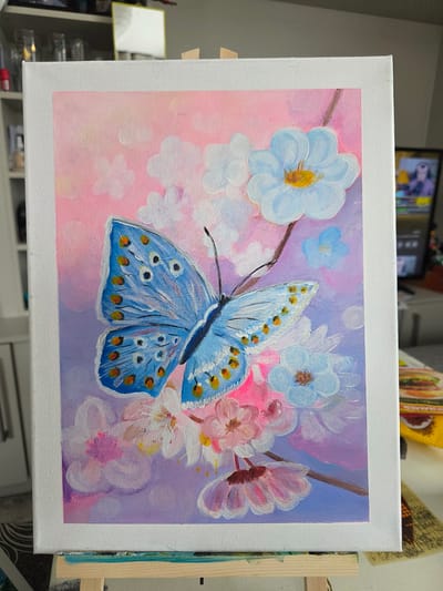 The image displays an acrylic painting of a blue butterfly amidst blossoms, positioned on an easel. The butterfly, with prominent orange-spotted wings, is centrally located. The blossoms vary in color, including white, pink, and light blue, with yellow centers on some. The background transitions from a pinkish hue at the top to shades of purple and blue at the bottom, suggesting a soft, atmospheric setting. The painting is on a white canvas, framed by the white border of the canvas. The easel is made of light-colored wood, with visible paint smudges on its surface. In the background, shelves are visible, holding various items including glass jars. A blurred television screen displaying an image of a person is also present. The scene appears to be indoors, possibly in a home studio or living space, and is not identifiable as being in Claremorris, Ireland, or any specific urban location. There are no people or interactions depicted in the painting itself.
