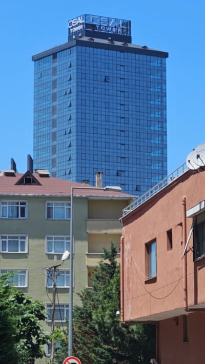 The image depicts an urban scene with a modern glass skyscraper juxtaposed against older, residential buildings. The skyscraper, identified by the text "OSAL TOWER" on its side, is clad in blue glass panels and stands tall against a clear blue sky. Several windows on the skyscraper are open, suggesting a warm day.

In the foreground, an older, salmon-colored building with a red-tiled roof features a satellite dish on its roof and a ladder extending along its side. Adjacent to it is a lighter green building with multiple windows, some of which have white frames. Trees with dense green foliage are visible around the buildings, adding a touch of nature to the urban landscape. A traffic sign with a red circle and a diagonal bar is partially visible on the right side of the image. The lighting suggests it is daytime, likely sunny, given the clear sky and the way light reflects off the buildings. The scene appears to be a typical urban environment, possibly in Ataşehir, Turkey, given the provided location context. There are no people visible in the frame.