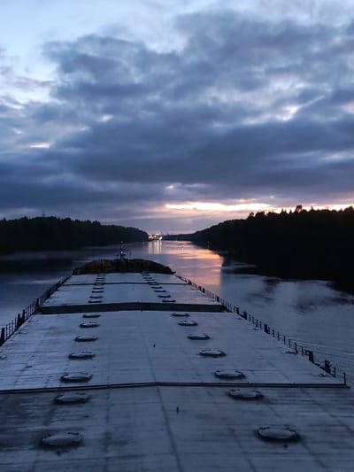 A low-angle photograph depicts a section of the Annensky Most bridge in Russia at dawn or dusk. The bridge appears to be a large, flat structure with a repetitive pattern of circular access points. Along the bridge's edges are short, vertical metal barriers. The river below reflects the ambient light and the illumination from distant lights on the shore. The banks of the river are lined with dark trees. The sky is dominated by a cloud cover with a narrow horizontal gap near the horizon, revealing the light from the setting or rising sun.