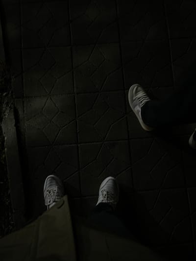 The scene depicts the feet of two individuals standing on dark, interlocking hexagonal pavement tiles in Karmana, Uzbekistan. In the lower foreground, a person wears light grey or white athletic shoes with white laces, partially obscured by the bottom edge of a light-colored garment, possibly a jacket or coat, hanging down. To the mid-right, a second individual's right foot is visible, wearing a similar light-colored athletic shoe with white laces and dark trousers. The environment is dimly lit, with subtle reflections on the textured tile surface, and a darker, unpaved area with minimal vegetation is present along the left edge of the frame. Both individuals appear to be standing still.