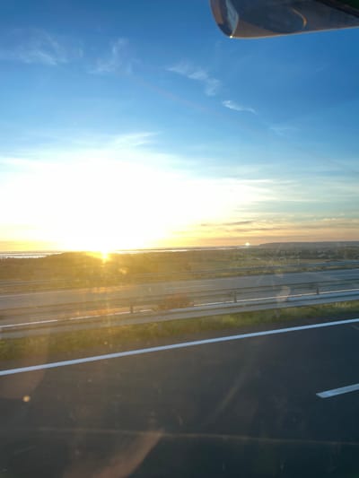 A wide-angle landscape photograph, taken from an elevated vantage point, depicts a sunrise or sunset over the region of La Palme, France. The upper sky displays a clear blue hue with scattered wispy clouds, transitioning into a bright yellow-orange near the horizon. The sun, a brilliant white orb, is positioned intensely on the left side of the horizon, generating significant glare and lens flare across the lower half of the sky and foreground.
Below the horizon, a large, flat body of water, consistent with the Étang de La Palme, is visible, reflecting the ambient light. In the mid-ground, a multi-lane highway, likely a section of the A9 autoroute, traverses horizontally. The highway features dark asphalt, clear white lane markings, and metallic crash barriers bordering a central reservation containing green grass and some low, reddish-brown vegetation. To the right of the highway, the distant landscape includes rolling hills with scattered structures, indicative of a settlement. A small, curved metallic component is visible in the top right corner of the frame, suggesting the image was captured from within a vehicle or aircraft. No individuals or specific actions are present.