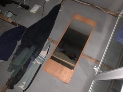 The image depicts a dimly lit room, possibly a storage area or a disused space, with a grey concrete floor and walls. In the foreground, a collection of clothing items is draped over a wooden beam. These include a dark navy garment, a green garment, and a garment with horizontal light and dark stripes. To the right, a tall, thin mirror with a wooden frame leans against the wall. The mirror reflects a person lying down, partially obscured by a blanket, and a portion of what appears to be a bed. In the background, a black electric guitar is visible on a surface, along with a small green notepad. A light blue mug or cup rests on a piece of wood in the mid-left of the frame. Electrical wires are strung along the top of the wall. The overall impression is one of a cluttered and somewhat neglected environment. The location is identified as Aksakovo, Russia.