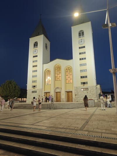 The image depicts the Church of St. James in Međugorje, Bosnia and Herzegovina, at dusk or night. The church features two prominent bell towers with clocks, illuminated by exterior lighting. The facade of the church is lit, revealing arched windows and large wooden doors. A group of people are gathered on the steps and in the plaza in front of the church, some are dressed casually and others in what appears to be religious attire. The plaza is paved with stone tiles, and a set of stone stairs leads up to the church entrance. A lamppost with an illuminated light is visible on the right side of the frame. The sky is dark blue, indicating it is nighttime or late dusk.