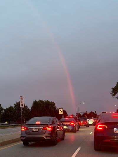 A faint rainbow arches across a hazy, overcast sky over a traffic jam on a street in Bloomingdale, United States of America. Several cars are stopped in multiple lanes, their brake lights illuminated. A "ONE WAY" sign with an arrow pointing right is visible on the left side of the road, as is another traffic sign indicating "ONLY" with a left-turn arrow. Trees line the sides of the road.