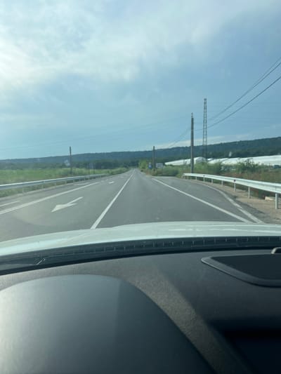 The image captures a forward-facing perspective from inside a vehicle on an asphalt road in the vicinity of Tsarkva, Bulgaria, under a partly cloudy sky. The road is a two-lane thoroughfare with white painted markings: a solid white line delineating the right shoulder, and a double white center line consisting of a solid line on the right and a dashed line on the left, indicating passing is permitted in the direction of travel. A large white arrow on the pavement in the left lane indicates the direction of travel is straight ahead.
To the left of the road, a silver metal guardrail runs parallel to the pavement. Beyond the guardrail, dense green vegetation borders the road, with a blue road sign mounted on a wooden utility pole, partially obscured and indistinct. Further down the road on the left, another white sign is visible on a pole. In the background on the left, a range of hills covered with dark green foliage extends horizontally.
To the right of the road, an identical silver metal guardrail is present, with patches of red flowers (likely poppies) growing along the embankment. Beyond the guardrail and vegetation, a series of large, white, tunnel-shaped agricultural structures, identifiable as greenhouses, are clearly visible, stretching into the distance. In the sky above the right side, multiple power lines are strung, supported by various utility poles, including a tall metal lattice tower and several wooden poles.
The sky occupies the upper portion of the frame, displaying scattered white clouds against a pale blue background. The lower portion of the image reveals the interior of the vehicle, specifically the dark gray dashboard with a textured surface and the lower edge of the windshield. No individuals or specific actions are discernible within the frame.