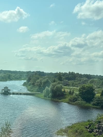 An aerial shot captures a wide river winding through a lush, green landscape under a partly cloudy sky. A small, docked structure, possibly a ferry terminal, sits on the left bank of the river, with a long pier extending into the water. The surrounding area is densely forested with various trees, and in the distance, hints of buildings and possibly agricultural fields are visible. The water's surface is rippled, suggesting a gentle breeze. A tiny airplane is visible in the upper right quadrant of the sky. The scene conveys a sense of natural tranquility and rural life, typical of the outskirts of a city like Mar'ino, Russia, known for its scenic rivers and natural beauty.