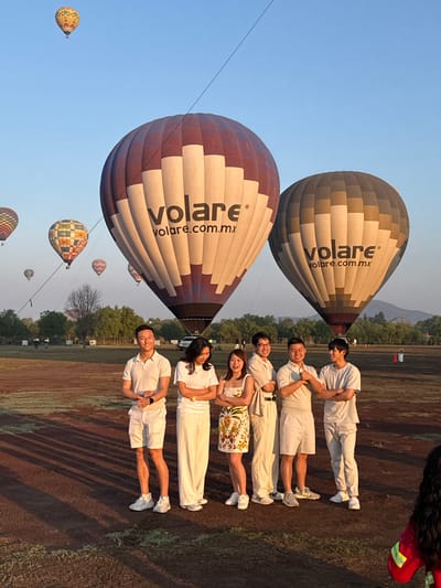 The image depicts a group of six people posing in front of two hot air balloons. The balloons, located in Ampliación San Francisco, Mexico, are labeled "volare.com.mx." The people are wearing casual clothing, mostly in light colors, and are lined up horizontally. Several other hot air balloons are visible in the background, floating in a clear sky. The ground appears to be a field with some sparse vegetation.