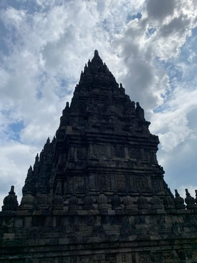 The image shows a low angle view of the Prambanan temple complex, located in the Kecamatan Prambanan district in Indonesia. The temple is a towering structure built of gray stone, with a series of tiers and intricate carvings. The top of the temple is obscured by a thick layer of clouds in the sky. There are no people visible in the image. The environment is a cloudy day.