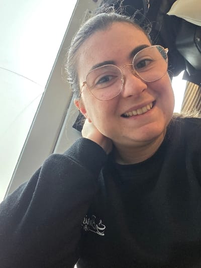 A female individual is depicted in a close-up portrait, smiling directly at the viewer. She has dark hair, wears round, gold-rimmed eyeglasses, and is dressed in a black long-sleeved top with a white stylized logo of text and a coffee cup design on the left chest. Her right hand is positioned with fingers resting near her jawline/neck. The background to the left features a bright, overexposed area, possibly a window, with a diagonal white structural element. The background to the right is darker and indistinct, showing textured material. The scene is located in the city of Gevgelija, North Macedonia.