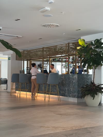 This media file shows the interior of a modern establishment, likely a bar or restaurant, with a sleek and contemporary design. The scene is well-lit, suggesting it is daytime. A prominent feature is a bar area with a marble-like countertop and a back bar filled with an assortment of bottles, indicating a selection of beverages.
The subjects are two individuals. One person, wearing a white shirt and beige pants, is standing behind the bar, seemingly engaged in preparing or serving something. Another person, wearing a light-colored shirt, is seated at the bar, facing away from the camera, suggesting they are a customer.
The setting is characterized by the use of light-colored marble flooring and dark, possibly blue, marble accents on the bar. There are several high stools with grey upholstery lined up at the bar. A large potted plant with broad green leaves adds a touch of nature to the sophisticated ambiance. The overall impression is one of a clean, upscale, and inviting space. The image does not provide explicit weather details, but the indoor lighting suggests controlled conditions. The location context indicates it is in Les Duanes De La Mar, Spain. There are no discernible emotions from the people depicted, as their faces are not clearly visible or are turned away. No legible text is visible within the frame.