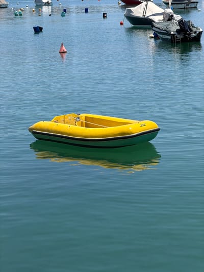 This media file shows a bright yellow inflatable boat floating on calm, blue water. The boat is positioned vertically in the frame and appears to be a RIB (Rigid Inflatable Boat) due to its rigid hull. It has a dark stripe running along its side and a rope attached to the front. The boat is casting a green reflection on the water's surface, which is slightly rippled, indicating a calm day.
In the background, to the left of the frame, several other boats are docked or moored. One boat is partially covered with a gray tarp, and another is white with a dark cover. Several buoys of various colors, including orange, blue, and white, are scattered in the water, marking the area. Some of these buoys appear to be small and spherical, while one orange buoy is cone-shaped with the letter "S" prominently displayed in black.
The overall scene suggests a marina or harbor environment, likely during daylight hours given the bright reflections on the water. The presence of multiple boats and buoys indicates a location where boats are kept or launched. The water's color suggests it's a coastal area or a large body of water, and the clear sky is implied by the bright lighting and reflections. There are no people visible in the frame. The scene evokes a sense of tranquility and stillness.