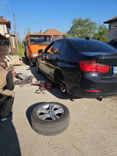 The image depicts an outdoor scene in Krasnovo, Bulgaria, on a bright, sunny day. A black BMW sedan is the central subject, elevated on a red hydraulic jack, with its rear passenger-side wheel removed. The corresponding wheel, featuring a silver BMW alloy rim and an all-season tire, rests on the concrete ground to the right of the car.
A person is observed from behind, crouching near the car and tools, indicating they are in the process of changing or repairing the tire. They are dressed in a beige jacket and dark pants. Numerous automotive tools are scattered on the ground, including a red and black cordless impact wrench, a comprehensive socket set in an open case, and various wrenches and small parts.
In the background, an orange van is parked close to the BMW. Further back, residential buildings with traditional terracotta roofs, green trees, and overhead power lines are visible against a clear, vibrant blue sky. The ground is a light-colored, textured concrete surface.
Notable details include the clear, sunny weather, indicating daytime. A partial view of the BMW's rear license plate shows "BG" (indicating Bulgaria) and parts of a number sequence "E D 4209". The detailed tread pattern of the all-season tire is clearly visible on the detached wheel. The overall setting suggests an informal vehicle maintenance activity taking place in a village or residential area.