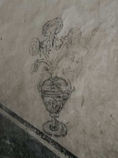 This image shows a black and white drawing on a stucco wall. The drawing depicts a vase of flowers. The vase is ornate with horizontal lines and a decorative base. The flowers above the vase are rendered with loose, sketchy lines. To the left of the vase, a dark, diagonal band runs across the bottom of the image, possibly representing a table or shelf. The wall itself appears aged and textured, with some faint smudges and discolorations. This drawing is located in the Convent of San Miguel Arcángel in Huejotzingo, Mexico.