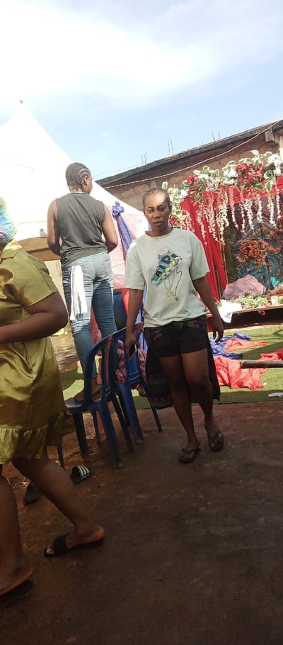 The image depicts an outdoor scene in Udi, Nigeria, under a bright, partly cloudy sky. The ground consists of dark brown, uneven packed earth or concrete.
Three individuals are visible. On the far left, a person's lower body and right arm are partially visible, wearing a golden or chartreuse-colored ruffled garment and black slip-on sandals. In the midground-left, a person with braided hair is facing away from the viewer, standing barefoot on a blue plastic chair. This individual wears a black sleeveless top and blue jeans with a white cloth piece tucked into the rear waistband. Their apparent action is interacting with a white tent structure or canopy positioned to their left. To the right of this person, another blue plastic chair is visible on the ground.
The central figure, a woman, faces forward with a neutral expression. She wears a light green short-sleeved t-shirt featuring a colorful sneaker graphic with an attached gold chain design. She also wears black shorts and a dark, patterned fabric or skirt tied around her waist/hips. Black flip-flops are on her feet, and a dark rectangular object, consistent with a mobile phone, is held in her right hand.
In the background, behind the central figures, a structure resembling a building wall is visible, with exposed rebar protruding from the top edge. Attached to this structure is an elaborate floral decoration, primarily featuring red and white flowers and green foliage, consistent with an event setup. Beneath this decoration, other scattered red and blue fabric pieces are on the ground, some resting on artificial green turf.