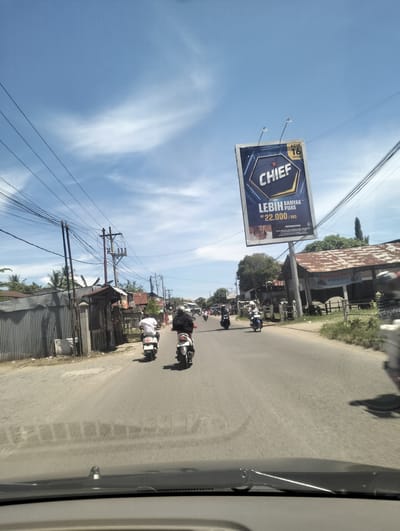 This media file captures a street scene in Miruektaman, Indonesia, on a sunny day. The sky is a bright blue with scattered white clouds, indicating good weather. The foreground shows the dashboard of a vehicle, suggesting the perspective is from a driver or passenger.
The main subjects are several people riding motorcycles on a paved road. Two motorcycles are clearly visible in the middle ground, with riders wearing helmets. Another motorcycle is further down the road, and more vehicles and riders are visible in the distance. The surrounding setting includes buildings with corrugated metal roofs, utility poles with a complex network of wires, and some vegetation. The overall atmosphere appears to be one of daily life and commuting.
A prominent object in the scene is a large billboard advertising "CHIEF". The billboard features text in Indonesian. Visible text on the billboard includes "NEW 16 BATANG", "CHIEF", "LEBIH PUAS", and "RP 22.000 / BKS". There's also a smaller section with "PERINGATAN" (Warning) and some illegible text below it. The billboard is positioned high on a pole, overlooking the road. The presence of the billboard suggests a commercial area or a road with significant traffic. The time of day appears to be daytime, likely late morning or early afternoon, given the bright sunlight and shadows.