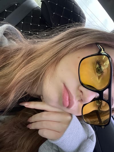 The image shows a close-up, low-angle shot of a young person lying down with their head resting on their hand, looking upwards. They are wearing a grey sweatshirt and large, black-framed sunglasses with yellow lenses. The sunglasses reflect the person's eye and the surrounding environment, which appears to be the interior of a vehicle, possibly a car, given the seat upholstery visible in the background. The person has long, light brown hair that is partially covering their face. The overall lighting suggests it is daytime, and the sky is visible through a window. The scene is likely taking place in or around the city of Новая Отрадовка, Russia, as specified by the user, although there are no explicit visual cues in the image to confirm this location.