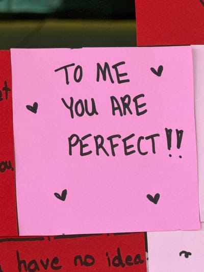 A square, pink sticky note displays a heartfelt message written in black marker. The text reads "TO ME YOU ARE PERFECT!!" and features small heart symbols above and below the words. The note is surrounded by other colorful sticky notes in red and white. A handwritten message "have no idea" is visible on a red note to the right. The image is casually captured, with uneven lighting and a slightly tilted perspective.