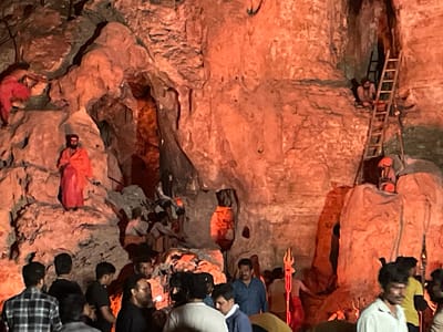The image depicts a lively gathering within a cave, possibly a religious site, in Gollagutta, India. The cave walls are a prominent feature, illuminated by warm, reddish light, giving the scene an otherworldly glow. Several individuals are visible throughout the cave, some appearing to be devotees or performers, while others are observers.
In the upper left portion of the image, a person in a red garment is positioned on a rocky ledge, seemingly in a contemplative or ritualistic posture. Below this, another individual, also in orange attire, is seated amongst the rocks.
Further into the cave, a wooden ladder is propped against the cave wall, leading to an upper recess. A person is seen seated near the top of the ladder, looking down. Another individual is positioned below the ladder, near the entrance of a darker crevice within the cave.
In the foreground and middle ground, a crowd of people are gathered, observing the activities or participating in the event. Many are looking towards the deeper parts of the cave. There is a prominent trident-like object standing vertically to the right of the center, suggesting a religious or spiritual context. The overall impression is one of a community event or pilgrimage taking place in a natural cave formation.