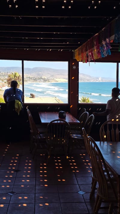 The image captures an interior view of a restaurant with a large window overlooking the ocean and coastline of Puerto Nuevo, Mexico. The foreground shows several wooden tables and chairs, with dappled light patterns cast on the tiled floor from an unseen light source, possibly decorative lights or sunlight filtered through a patterned structure.
Two individuals are visible. On the left, a person with dark hair pulled back is leaning against a dark surface, looking out the window. They are wearing a blue shirt and a yellow cloth is draped over their shoulder. On the right, a person facing away from the camera is standing near the window, also looking out at the sea. They are wearing a light-colored polo shirt.
The window reveals a scenic view of the Pacific Ocean with waves breaking on the shore. The coastline is dotted with buildings, and hills rise in the background under a bright sky. Colorful papel picado decorations are strung across the ceiling beams. The overall impression is that of a casual dining establishment with a prime ocean view.