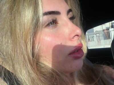 This is a close-up, profile shot of a young Caucasian woman with blonde hair, looking to her right. The image is taken outdoors, with a blurred background suggesting an urban environment. Sunlight casts a shadow across her face, highlighting her features. She has full, plump lips, applied with a glossy, pinkish-red lipstick. Her eyebrows are dark and well-defined, and she appears to be wearing false eyelashes. A small mole is visible on her cheek. The backdrop shows a white fence and some buildings, possibly residential or commercial, with a clear sky above. The setting is consistent with an outdoor location in Sant Vicent del Raspeig, Spain, during daylight hours.