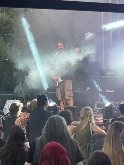 The image depicts a live outdoor music performance at the Analiontas Festival in Analiontas, Cyprus. A male musician, wearing khaki shorts and a dark t-shirt, is on stage playing an electric guitar. He is surrounded by theatrical smoke and illuminated by a bright spotlight. Behind him, two large "Marshall" brand amplifier cabinets are visible. The audience is gathered in front of the stage, with several individuals visible in the foreground, facing the performer. The background includes trees and lighting rigs, indicating an outdoor venue.