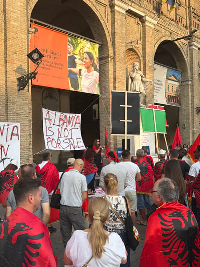 The image depicts a street protest in Parma, Italy, in front of a brick building with arched entrances. In the foreground, a crowd of people, many wearing red capes with the black double-headed eagle symbol of Albania, are gathered. They are holding banners. One prominent banner reads "ALBANIA IS NOT FOR SALE" in large, red, handwritten letters. Another banner, partially visible on the left, also features the Albanian symbol. Behind the protestors, a statue of a man in a toga stands on a pedestal. Several large banners are hung on the building's facade. One, orange, advertises a program called "Nasce il Coach Quartiere" from "Parma". Another banner, partially visible to the right of the statue, has text and an image of a building, likely related to the "Centro Centro Famiglie" of "Municipio". The scene appears to be outdoors during daylight, with sunlight casting shadows. A woman on the right side of the image is speaking into a microphone.