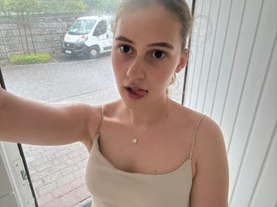 A young woman is taking a selfie in Forbach, Germany. She is wearing a light-colored tank top and a heart-shaped pendant necklace. Her face is close to the camera, and she is sticking out her tongue. Her eyes are wide open, and her eyebrows are raised. Her hair is pulled back in a bun.
The background of the image shows a white wall with vertical slats on the left and a window on the right. Outside the window, it appears to be raining heavily. There is a white van visible in the background, which is driving on a street with brick pavement. The weather appears to be overcast, and the time of day is not specified.
The overall mood of the image is playful and lighthearted, despite the inclement weather. The woman's expression suggests a sense of fun and mischief.