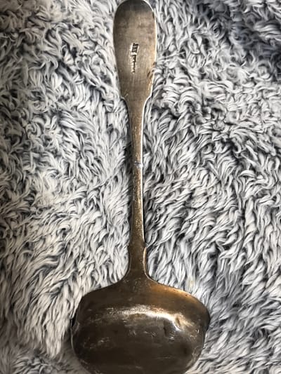 A silver ladle rests on a grey, fuzzy blanket. The ladle has a long, slender handle with a hallmark stamped on its upper portion. The bowl of the ladle is wide and shallow, showing signs of age and use. The texture of the blanket is soft and shaggy, with individual strands clearly visible. The lighting in the image is soft, creating subtle shadows and highlights on the metallic surface of the ladle. There are no people present in the image, and the focus is entirely on the ladle and the blanket. The location in Ново-Талицы, Russia, is not discernible from the image itself, as there are no contextual clues to indicate this specific geographical setting.