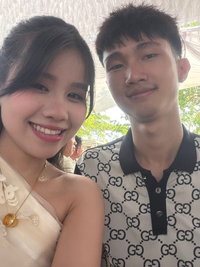 A close-up, upward-angled selfie captures a smiling man and woman, likely a couple, at an outdoor or semi-outdoor event in Bình An, Vietnam. The image is oriented vertically, with the man positioned towards the upper left and the woman in the foreground towards the lower right, both looking towards the camera with warm expressions.

The man has short dark hair and a small earring in his left ear. He is wearing a black and white polo shirt patterned with prominent "GG" logos, featuring a black collar and a placket with two visible silver buttons.

The woman has long dark hair and a wide, bright smile, revealing her teeth. She is wearing a light-colored, possibly cream or white, dress or top with subtle lacy embellishments. She is adorned with a delicate gold necklace featuring a small pendant and a gold ring on her finger.

In the blurred background, green foliage and bright natural light suggest a sunny daytime setting, likely under a white tent or canopy, parts of which are visible in the upper left. Another person's out-of-focus head is faintly visible behind the woman. The overall mood conveyed by the subjects' smiles is joyful and celebratory.
