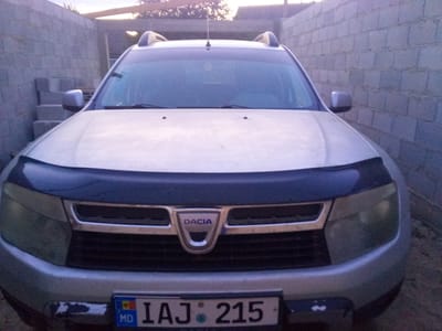 The image shows the front of a silver Dacia Duster parked outdoors, likely in a residential or industrial area in Leușeni, Moldova. The car's license plate reads "IAJ 215" with the MD country code. The car is facing the camera, and its headlights and grille are clearly visible. The background consists of a brick wall and some construction materials. The sky is visible at the top of the image, indicating it is daytime. The overall scene appears to be a still shot of a parked vehicle.