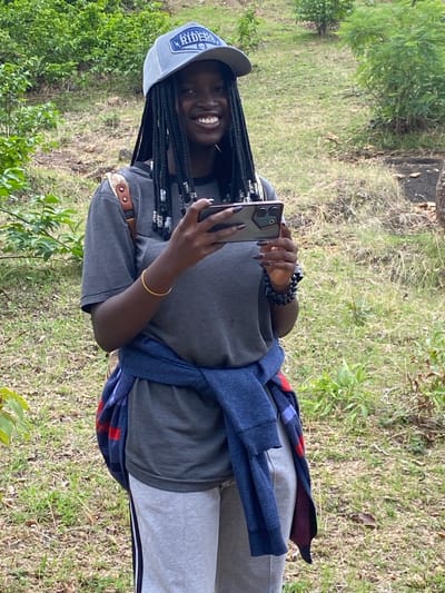 A person is standing in a grassy, wooded area, holding a smartphone in both hands and taking a photograph. The person is wearing a gray t-shirt, light gray sweatpants, and a gray baseball cap with "RIDERS" printed on it. A blue jacket is tied around their waist. They have long braids with decorative silver beads. A gold bracelet is visible on their right wrist. The setting appears to be outdoors in a natural environment with green foliage and grass. The location is Ganawuri, Nigeria.