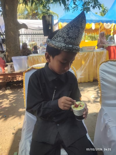 A young boy, likely of Southeast Asian descent, is the central subject of the image, seated and engrossed in eating from a small cup. He is wearing a distinctive, ornate conical hat with black and white patterns, often referred to as a songkok or a similar traditional headwear. His attire consists of a long-sleeved black shirt with buttons down the front and black trousers, suggesting a formal or semi-formal occasion. He holds a small white paper cup with a grid pattern, which appears to contain a light-colored dessert, possibly ice cream or shaved ice, topped with sprinkles and a red fruit piece. He is using a small red spoon to eat.

The setting is an outdoor event under bright daylight, indicated by the strong shadows and clear blue sky visible through tree leaves. The boy is sitting on a white chair, likely covered with a white chair cover, typical for outdoor events. In the background, there are several white and yellow tents or canopies, suggesting a festive gathering. Tables covered with yellow fabric tablecloths are visible, adorned with various dishes or serving bowls, including a white bowl and a red container in the distance.

Other people are present in the background; to the far left, a woman wearing a light brown hijab is partially visible sitting behind a white fence, under the shade of a large tree trunk. Further back, another woman in a light brown hijab and a woman in a white hijab are seen near the yellow-covered tables. A black speaker is mounted on a stand or pole near the tree. A rustic wooden table with intricate carvings holds a large, clear plastic container on the left side of the frame. The ground appears to be a mix of dirt and scattered leaves.

In the bottom right corner of the image, a timestamp reads "05/02/2026 14:31", indicating the date and time the photograph was taken. The overall atmosphere appears to be relaxed and celebratory, typical of a family or community gathering.