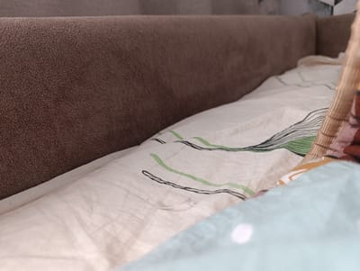 The image depicts a close-up, low-angle view of a bed within an indoor setting, potentially a bedroom. The dominant element is a brown, textured headboard that runs horizontally across the left and upper portions of the frame. Beneath the headboard, a light-colored duvet or comforter is visible, featuring a pattern of thin, wavy lines in black and green. A white thread is also present on the duvet. In the foreground, a portion of a light blue blanket with white circular polka dots is visible. The overall impression is of a domestic environment. The image was taken in Krasnyi Sad, Russia.