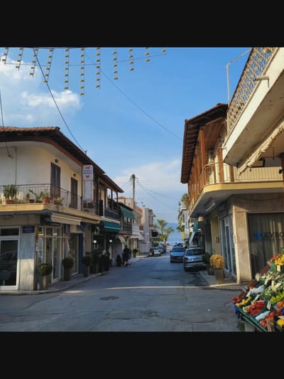 The image depicts a street scene with buildings lining both sides of a paved road. The sky is blue with scattered clouds. String lights are visible overhead, spanning across the street. On the left side of the street, a building with a balcony adorned with potted plants is prominent. A sign with Greek text and a graphic of a pig is affixed to the building. Further down the street, on the right side, a building with a wooden balcony and railing is visible. A fruit stand is set up on the sidewalk in the foreground on the right, displaying a variety of colorful fruits and vegetables. Several cars are parked along the street. A few people are present, walking or standing. The overall impression is of a somewhat quiet street in a European town, possibly a coastal area, given the distant view of water. The location provided, Bielawska Wieś, Poland, is inconsistent with the Greek signage in the image, suggesting a potential error in the location data.
