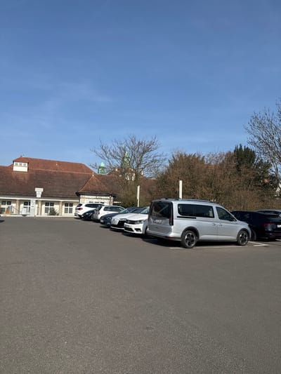 The image captures an outdoor asphalt parking lot in Bad Nauheim, Germany, under a clear, light blue sky. The scene is illuminated by direct sunlight. In the midground, a row of approximately eight passenger vehicles is parked, facing away from the viewer. The most prominent vehicle is a silver (light grey) Volkswagen Caddy-style van, parked perpendicular to the camera, displaying its rear and left side and a German "FB" license plate. To its left, a white Volkswagen, a dark Audi sedan, another white car, a dark SUV, and a white SUV are visible. A dark SUV is partially visible to the right of the silver van. Behind the parked vehicles, a line of deciduous trees and bushes, largely bare, delineate the property line. Two light-colored vertical poles rise above the foliage. In the background, to the left, a multi-story building with a distinctive reddish-brown tiled roof and a white facade on its lower levels is visible, consistent with the architecture of the Sprudelhof complex. A small evergreen tree is positioned near the building's base, and a grey payment kiosk stands on the far left. Further behind the trees, a green-domed structure, characteristic of the Sprudelhof's central building or a nearby church, is partially visible. No people are present.