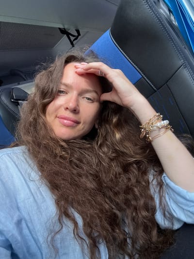 A woman with long, curly brown hair and fair skin is taking a selfie inside what appears to be a vehicle. She is wearing a light blue linen shirt and several gold bracelets with pearls on her right wrist. Her right hand is resting on her forehead. The background shows dark upholstery with blue trim, consistent with seating in a bus or train. The lighting suggests it is daytime, possibly indoors or in a shaded area. There is no visible indication of the location being in the city of Lisava, Russia.