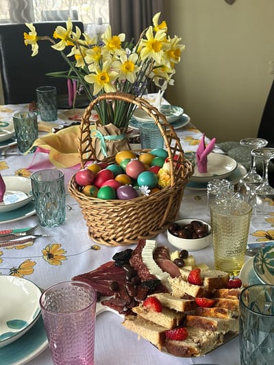 A table is set with a flowered tablecloth, plates, glasses, silverware, napkins, and a basket of Easter eggs. There is also a platter of food including bread, cheese, strawberries, and cured meats, a small bowl of olives, and a vase of yellow flowers. The location is Bryastovets, Bulgaria.