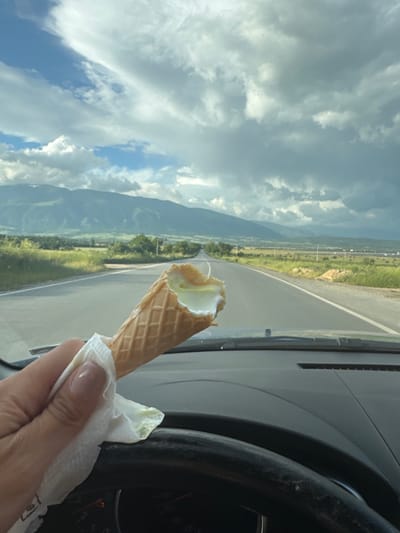 An ice cream cone with a bite taken out is being held in a hand inside a car. The background shows a road and landscape with mountains, visible through the car's windshield. The car's steering wheel is also visible. The location appears to be in or near Vedrare, Bulgaria, based on landscape characteristics.