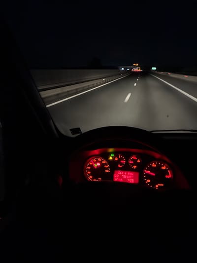 The image captures a night-time view from inside a vehicle driving on a highway. The dashboard lights, emitting a red glow, are prominently visible in the foreground, displaying the speedometer, fuel gauge, and an LCD screen. The LCD screen shows a mileage of 759900. The highway stretches ahead, illuminated by the vehicle's headlights and distant streetlights, with lane markings clearly visible. The overall scene suggests a journey on a deserted highway, possibly in the vicinity of Remetea Mare, Romania, given the context. There are no people or animals visible in the image.