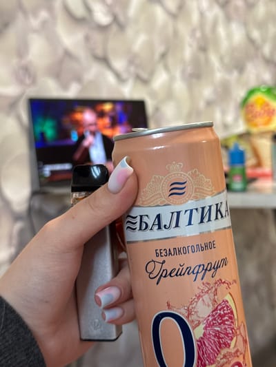 An indoor scene in Shirkovo, Russia, shows a person's hand holding two objects. The hand, with light skin and partially visible in a dark grey sleeve, has fingernails styled with a light base and soft pink and blue accents. In the left grip is a metallic grey rectangular device with a black top, likely a vape, featuring a subtle "V" logo near its base. To the right, the hand firmly grasps an aluminum can of Baltika 0% Grapefruit non-alcoholic beer. The can is peach-orange, displaying the brand "БАЛТИКА" (BALTIKA), the text "БЕЗАЛКОГОЛЬНОЕ" (NON-ALCOHOLIC) and "Грейпфрут" (Grapefruit), along with a large "O" and a graphic of a grapefruit slice with splashes. The can's upper section features a crowned emblem indicating "BREWING COMPANY BALTIKA" and "FOUNDED IN 1990." In the blurred background, a television screen displays a vibrant, colorful performance with a faint male figure holding a microphone. The wall behind is covered with a light-toned, subtly patterned wallpaper. To the right of the screen, out of focus, are a green and yellow object and a small blue bottle with a green cap on a surface.