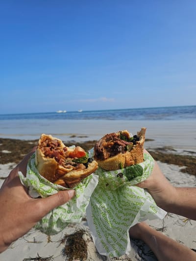A sideways, medium shot depicts two hands holding half-eaten sandwiches wrapped in white paper printed with green lettering, against a background of a beach and a clear blue sky. The sandwiches appear to be filled with meat, vegetables, and olives. The beach has white sand and brown seaweed. The hands holding the sandwiches are those of a fair-skinned person, possibly two people. The sandwiches obscure the person's body, leaving only their hands and feet visible. The location is Big Pine Key, United States.