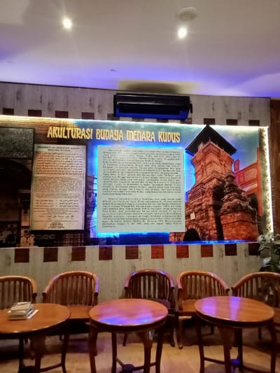 The image depicts a scene inside a restaurant or cafe located in Kudus, Indonesia, at coordinates -6.809, 110.843. The interior features wooden walls with a brick pattern and a large, framed image of a historical tower. The image, titled "AKULTURASI BUDAYA MENARA KUDUS," includes a caption describing the historical and cultural significance of the tower. There is a display case with artifacts or exhibits behind the table. The room is dimly lit by overhead lights. Four wooden chairs are situated around two tables, with one table empty and the other table containing several books. The chairs are mostly empty. One chair has a wooden slatted back and another chair has a solid wooden back. There is a potted plant in the corner. The scene is empty of people, conveying an atmosphere of stillness.